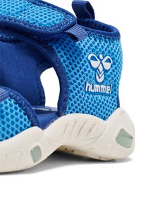 Niebieski siateczkowy but sportowy z rzepem, teksturowana powierzchnia i biała gumowa podeszwa. Zawiera wyraźne logo po boku.