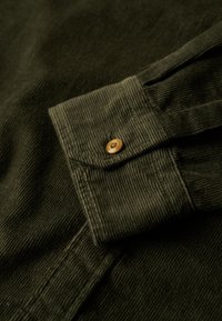 Olive green corduroy shirt detail featuring a buttoned cuff and textured fabric with vertical stripes. Natural wooden button accent.