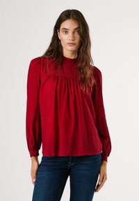 Blusa rossa con texture, colletto alto, dettagli plissettati e maniche lunghe. Abbinata a jeans blu scuro ne esalta il contrasto di colore.
