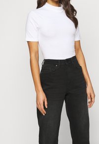 Kvinna i en åtsittande vit kortärmad topp med hög krage, instoppad i högmidjade svarta jeans med framfickor och bälteshällor.