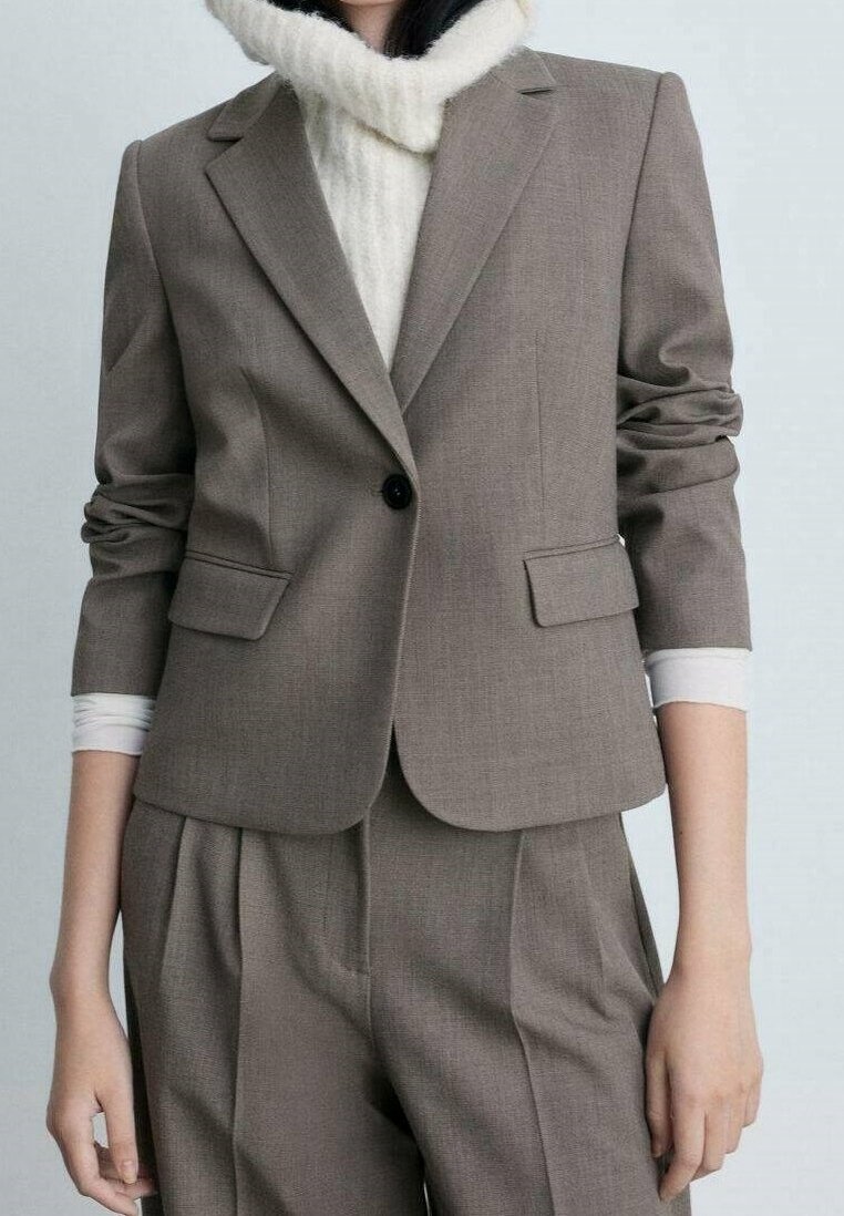 Femme portant un blazer gris sur mesure par-dessus un pull à col roulé blanc avec un pantalon plissé gris assorti.