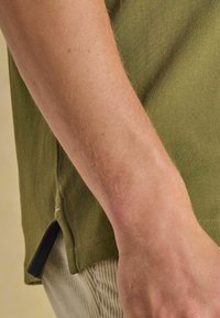 Olivengrünes Kurzarmhemd aus strukturiertem Stoff, mit Seitenschlitzen und einem schwarzen Akzent am Saum, kombiniert mit hellen Hosen.