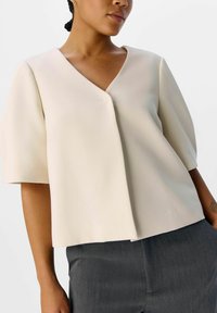 T-shirt crème à manches courtes avec un col en V et une coupe décontractée. Fabriqué en tissu lisse, il présente un design à l'avant légèrement superposé.