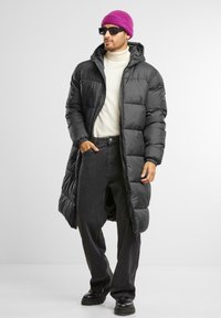 Zwarte pufferjas met een lange uitstraling en capuchon, gedragen over een witte coltrui. Zwarte jeans en chunky zwarte laarzen maken de outfits compleet.