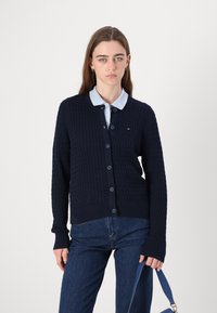 Σκούρα μπλε πλεκτή ζακέτα με υφαντό σχέδιο, στρογγυλή λαιμόκοψη και κουμπιά μπροστά. Φοριέται πάνω από ένα ανοιχτό μπλε πουκάμισο με γιακά, συνδυασμένο με τζιν.