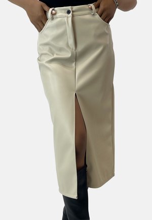 Beige midi-nederdel med høj talje, foran med slidser, bæltestropper og knap, båret med sorte knæhøje støvler og hænder i lommerne.