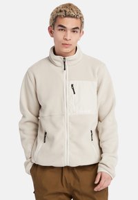 Lys beige fleecejakke med høj krave, lynlås foran og brystlomme. Har sorte lynlåsdetaljer og et logo på lommen.