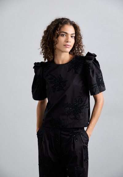 Haut noir à manches courtes avec un tissu texturé, des embellissements en forme de feuilles, des accents perlés et des épaules bouffantes structurées.