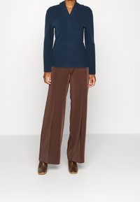Pull-over en maille bleu marine à col en V, associé à un pantalon large marron et des bottines marron à talons, avec des textures lisses et des lignes épurées.
