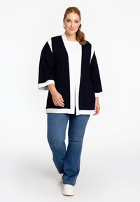 Marineblauer Cardigan mit weißen Akzenten, offenes Frontdesign, 3/4-Ärmel, kombiniert mit blauen Jeans und weißen Sneakers, glatte Stofftextur.