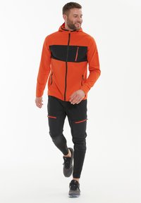 Orangene und schwarze kapuzensichere Jacke mit einem schwarzen Vorderteil, mit Reißverschlusstaschen, und schwarzen, taillierten Hosen mit roten Akzenten.
