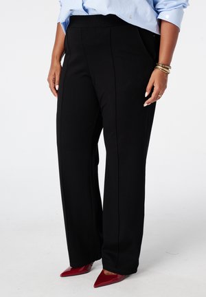 Personne portant un pantalon noir à jambes larges, une chemise bleu clair, des chaussures rouges pointues et des bracelets en or au poignet gauche sur fond blanc.