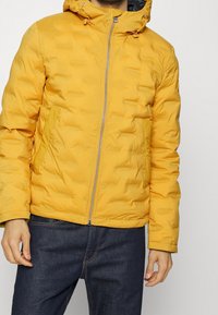Gul pufferjacka med huva, med dragkedjestängning, texturerad yta och sidofickor, kombinerad med mörka jeans.