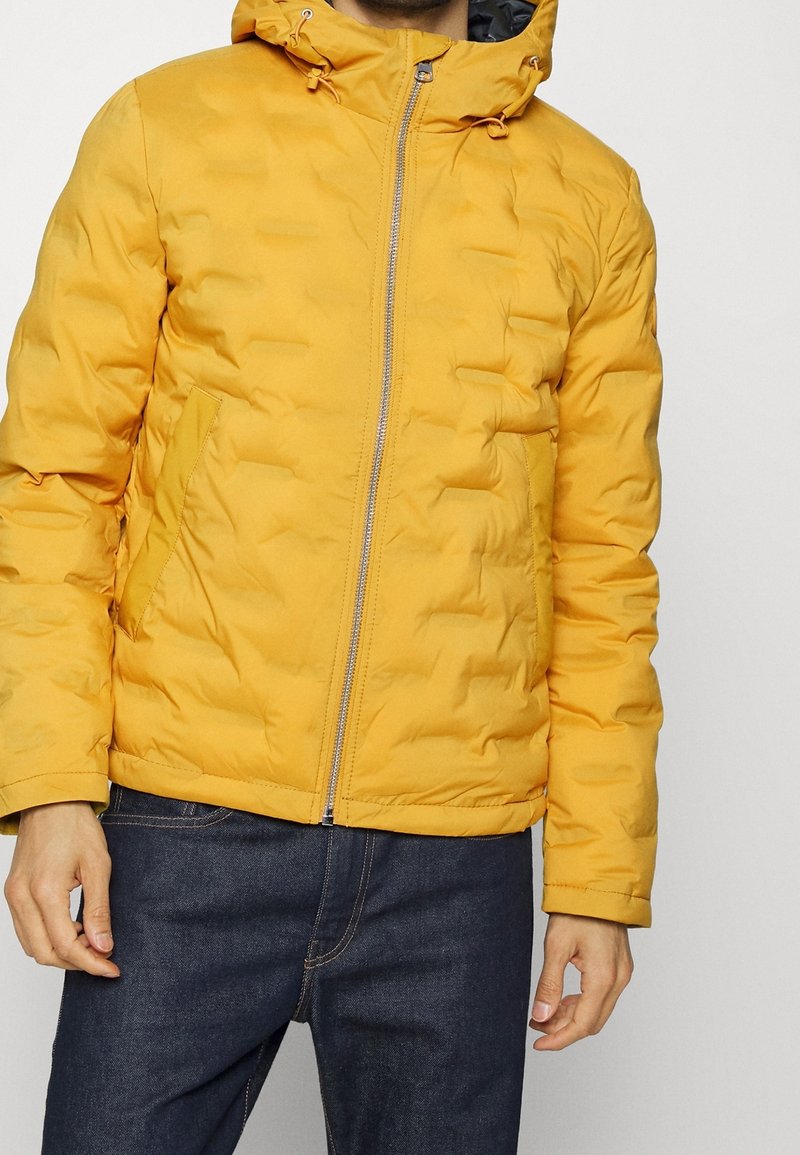 Gul pufferjacka med huva, med dragkedjestängning, texturerad yta och sidofickor, kombinerad med mörka jeans.