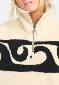 Pullover in pile, crema con design astratto a vortice nero. Presenta un colletto alto e una cerniera frontale con hardware metallico. Texture morbida.