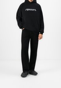 Personne portant un sweat à capuche Rip Curl noir, un pantalon noir et des baskets noires, debout devant un fond blanc uni.