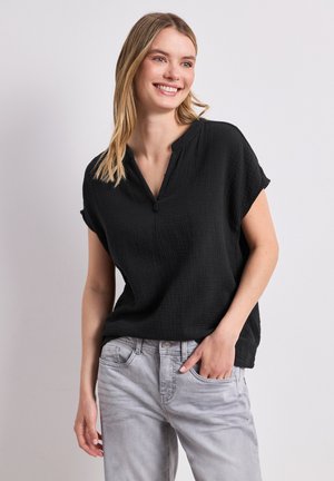 Uśmiechnięta kobieta o długich blond włosach, ubrana w czarną bluzkę z krótkim rękawem i jasnoszare dżinsy, pozująca z jedną ręką w kieszeni.