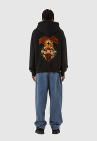 Felpa nera con grafica sul retro che presenta un cuore, rose e fiamme. Grande testo rosso recita "migueltales." Indossata con pantaloni in denim blu e scarpe nere.