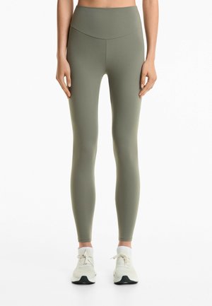 Leggings taille haute vert olive en tissu extensible, présentant une texture lisse et un design ajusté. Associés à des chaussures de sport légères.