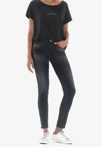 Czarny t-shirt z krótkim rękawem z złotym napisem, zestawiony z dopasowanymi ciemnoszarymi dżinsami. Modelka w białych trampkach, stojąca na jednolitym tle.