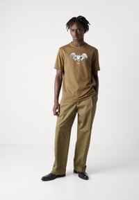Brown cotton t-shirt featuring a bat graphic and the word "BOSS" below it. Paired with olive green trousers and black shoes.