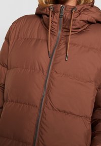 Brun vadderad jacka med dragkedja fram, justerbar huva och grå dragkedjor. Har kviltade sektioner och en slät yta.