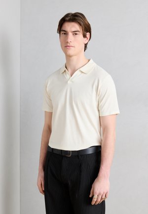 Ung mann iført en kremfarget kortermet polo-skjorte som er stukket inn i svarte pinstripe-bukser med svart belte, stående mot en ensfarget vegg.