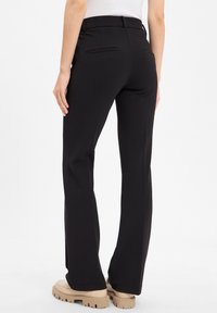 Schwarze weit geschnittene Hosen mit glatter Textur, versehen mit Gesäßtaschen und einem taillierten Bund. Sichtbar sind beigen Schuhe mit dickem Absatz.