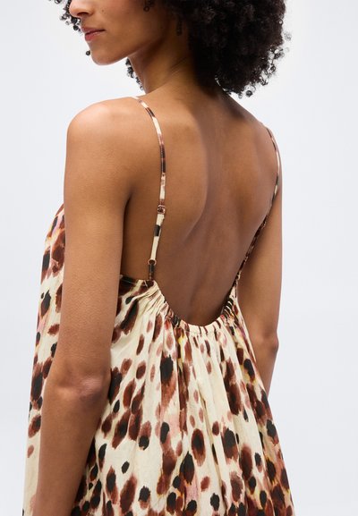 Γυναίκα με σγουρά μαλλιά που φοράει αμάνικο φόρεμα με λεοπάρ σχέδιο, λεπτές ρυθμιζόμενες τιράντες και ανοιχτή, χαμηλή πλάτη.