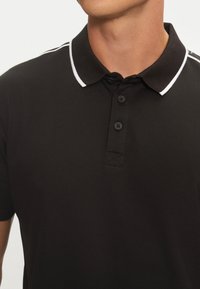 Camisa polo negra con cuello clásico, que presenta un ribete de acento blanco, tres botones negros y una tela texturizada.
