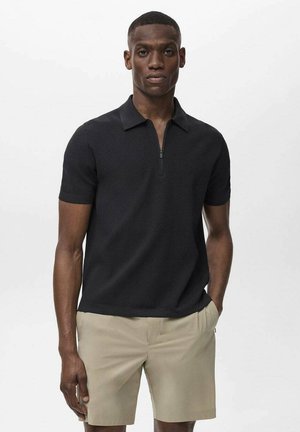 Polo shirt nero a maniche corte con colletto zip e texture a coste, abbinato a pantaloni beige. Design minimalista, senza motivi o dettagli.