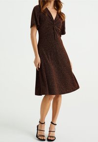 Brun, glittrig knälång klänning med korta, svävande ärmar och en vriden frontdetalj, tillverkad av ett mjukt, strukturerat tyg. Svarta remsandaler.