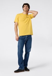 Junger Mann, der mit einer Hand hinter dem Nacken steht, in einem gelben kurzärmeligen T-Shirt, blauen Jeans und schwarzen Turnschuhen vor einem weißen Hintergrund.