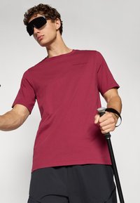 Man wearing black sunglasses and a red t-shirt holding a black hiking pole with gray handle against a plain background.