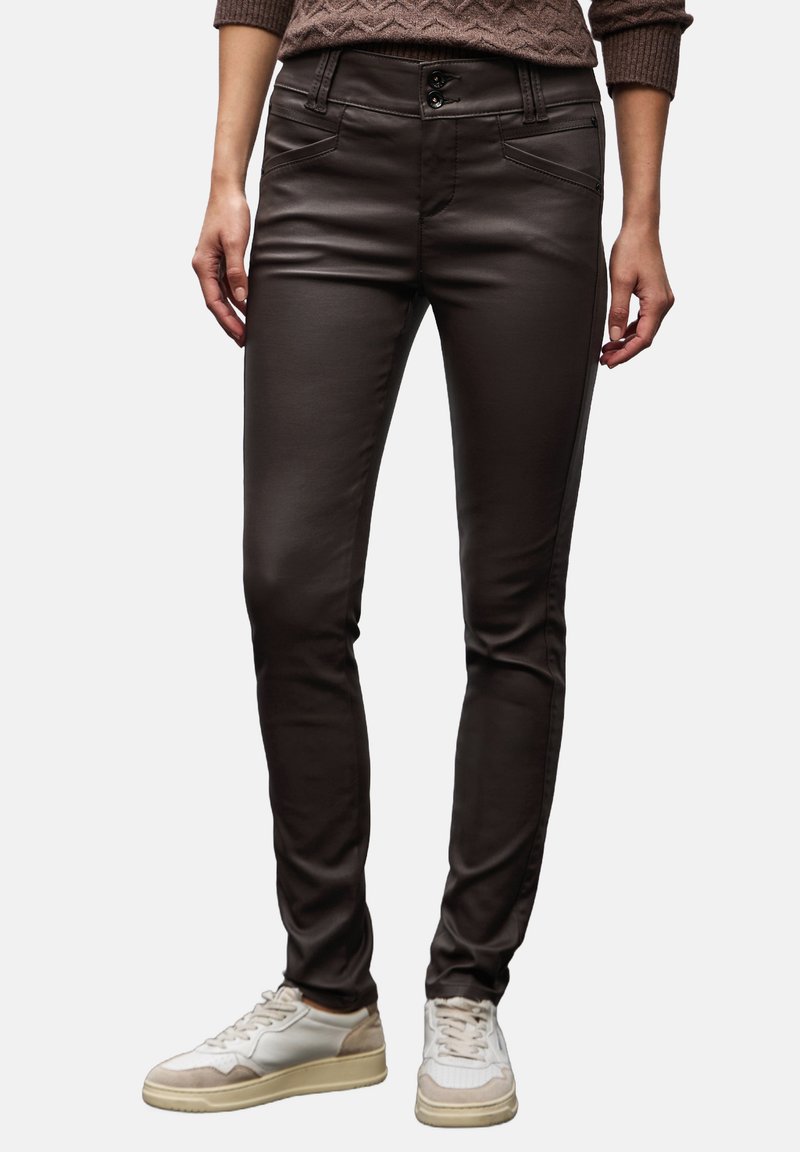 Pantalones ajustados de color marrón oscuro, confeccionados con una tela lisa y elástica, que cuentan con dos bolsillos frontales y un cierre de doble botón. Llevados con zapatillas blancas.
