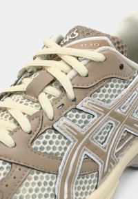 Zapatilla deportiva beige con parte superior de malla y suela de goma. Presenta detalles en un tono beige claro contrastante, cordones redondos y detalles de la marca en la lengüeta.