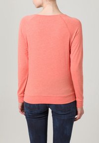 Korálový pulovr z měkké látky, s dlouhými rukávy, kulatým výstřihem a volným střihem, doplněný tmavě modrými džínami.