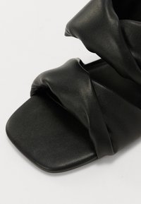 Sandali in pelle nera con punta quadrata, caratterizzati da fasce superiori attorcigliate e una texture liscia. Il design è minimalista e contemporaneo.
