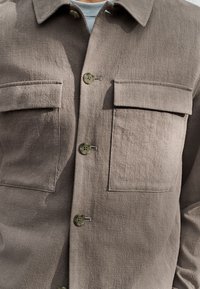 Camisa marrón abotonada hecha de tela texturizada, con dos bolsillos en el pecho, botones verdes y cuello con solapa.
