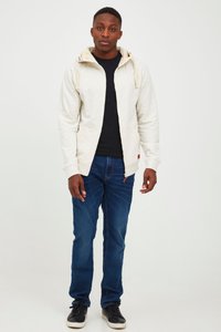 Svetlošedá mikina na zips s kapucňou so sťahovacou šnúrkou, nosená cez čierne tričko. Modré rifle a čierne topánky dopĺňajú outfit.