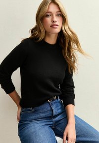 Schwarzer Strickpullover mit langen Ärmeln und rundem Hals, kombiniert mit hochtaillierten blauen Jeans. Weicher Stoff, figurbetonter Schnitt.