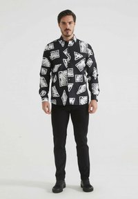 Chemise noire avec de grands motifs géométriques abstraits blancs, comprenant des triangles et des rectangles. Manches longues, design à boutons, en coton.