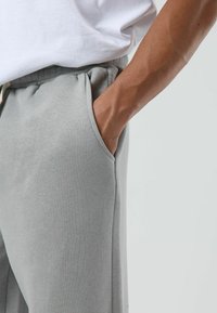 Pantalon de jogging gris clair avec une taille élastique et des poches latérales, présentant une texture lisse et une coupe décontractée, montré à côté d'une chemise blanche.