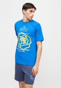 Νέος άνδρας που φοράει μπλε μπλουζάκι με γραφικά NYC και γκρι σορτς New Balance, κοιτάζοντας προς την πλευρά σε απλό φόντο.