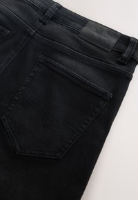 Schwarze Jeans aus Denim, gefaltet, so dass die Gesäßtasche und der Bund mit einem schwarzen Leder-Branddetail sichtbar sind.