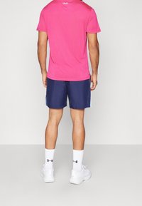 Man som står med ryggen vänd, iförd en rosa kortärmad skjorta, marinblå shorts med ljusblå sidostråkar, vita strumpor och vita sneakers.