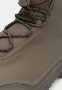 Nærbilde av beige fjellsko med tykk såle, mønstrede snører og "STOP EXPLORING" stropp med The North Face-logoen.
