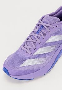 Zapatilla deportiva morada con parte superior de malla texturada, acentos de rayas blancas y un cuello acolchado. La suela azul claro completa el diseño.