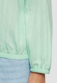 Blouse vert menthe avec un tissu texturé et froissé. Équipée de poignets élastiques et d'un ourlet arrondi, associée à un denim bleu clair.