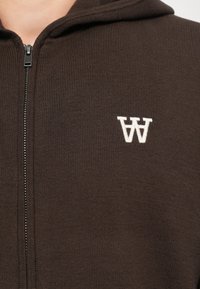 Brown zip-up hoodie made from ribbed fabric, featuring a white logo on the chest, and a textured, soft finish.